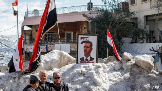Druze residents of the Golan Heights stand by flags of Syria and a portrait of Syrian President Bashar al-Assad fixed in a mound of shoveled snow at a square by a monument commemorating the 1925 Battle of al-Mazraa in the Druze revolt against France, in the Druze village of Majdal Shams in the Israeli-annexed Golan Heights on February 14, 2023, as locals gather for a rally against the 1981 Israeli annexation law of the strategic plateau which the Jewish state captured from Syria during the 1967 Arab-Israeli war. (Photo by JALAA MAREY / AFP)