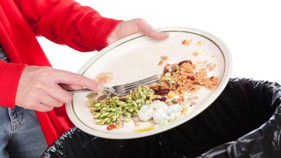 Close-up of a woman sweeping the leftovers from a meal into a domestic garbage bin. The background is pure white.