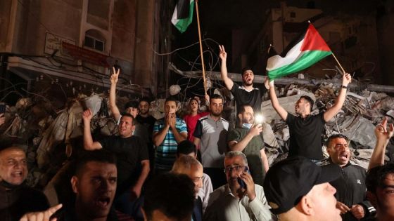 A man waves the Palestinian flag as others flash the V-sign for victory as they celebrates in front of a destroyed building the ceasefire brokered by Egypt between Israel and the two main Palestinian armed groups in Gaza on May 20, 2021. - Israel and the two main Palestinian armed groups in Gaza, Hamas and Islamic Jihad, announced a ceasefire on May 20, 2021, aimed to end the most devastating conflict between them for seven years. The truce brokered by Egypt was announced following mounting international pressure to end 11 days of conflict that has claimed lives on both sides, with Israeli jets pounding Gaza with air strikes as militants fired thousands of rockets towards Israel. (Photo by MOHAMMED ABED / AFP)
