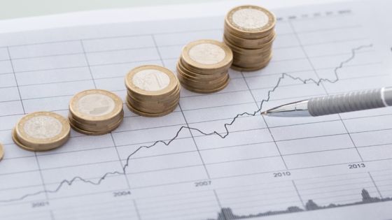 Cropped image of businessman's hand with pen analyzing line and coins graphs on desk