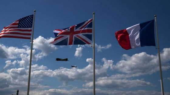 National flags from US, United Kingdom and France fly in foreground as a Douglass C-47 Dakota (L) and a Sptifire fly over Arromanches-les-Bains, northwestern France, on June 6, 2019, as part of D-Day commemorations marking the 75th anniversary of the World War II Allied landings in Normandy. (Photo by JOEL SAGET / AFP)