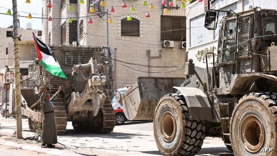 A Palestinian man waves the Palestine flag as Israeli army vehicles block the entrance to the Tulkarem camp for Palestinian refugees, in the occupied Palestinian West Bank on August 22, 2024. (Photo by Zain JAAFAR / AFP)