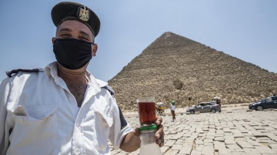 A mask-clad Egyptian policeman on duty at the Giza Pyramids necropolis walks while holding a glass of tea and a jar of sugar past the Great Pyramid of Khufu (Cheops), open for tourists visiting the archaeological site on the southwestern outskirts of the Egyptian capital Cairo on July 1, 2020 as the country eases restrictions put in place due to the COVID-19 coronavirus pandemic. - A spree of openings in Egypt comes after the country officially ended a three-month nighttime curfew a few days earlier. Cafes and shops have re-opened but public beaches and parks remain closed as part of measures to curb the spread of the novel coronavirus. Egypt has recorded more than 65,000 COVID-19 cases including over 2,700 deaths. (Photo by Khaled DESOUKI / AFP) (Photo by KHALED DESOUKI/AFP via Getty Images)