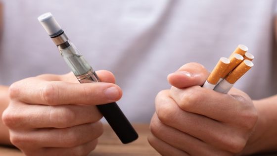 Man Holding Vape And Tobacco Cigarette Over Desk