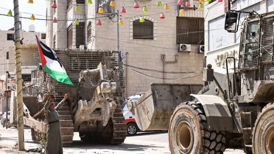 A Palestinian man waves the Palestine flag as Israeli army vehicles block the entrance to the Tulkarem camp for Palestinian refugees, in the occupied Palestinian West Bank on August 22, 2024. (Photo by Zain JAAFAR / AFP)